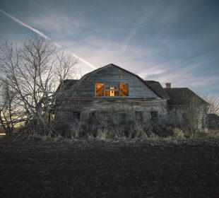 A photograph of the eastern side of an old and sun-faded wooden house. The windows are missing their panes and the wood is gray with age. There are leafless trees, seemingly because of it being early spring or late winter when the photo was taken, along the edge of the building and a light seems to be glowing from inside the house. The sun is setting on the other side of the building and some stars are twinkling in the lightly clouded blue sky above the building. 