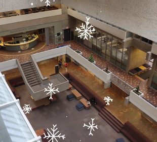 The Administration Humanities conversation pit is pictured from the fifth floor of the building. White snowflakes are drawn overtop the photo and are falling into the pit from the fifth floor. 