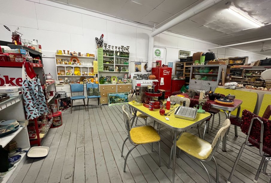 An image of an antique store, there are many different sorts of wares for sale. There is a Coke-branded fridge in the background; a few tables and chairs covered in other things; there’s a painting propped up on the floor near the back; martini glasses, milk bottles, and other dishes on a shelf at the back; and many other things that aren’t particularly discernable scattered throughout the room.