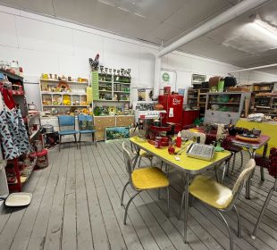 An image of an antique store, there are many different sorts of wares for sale. There is a Coke-branded fridge in the background; a few tables and chairs covered in other things; there’s a painting propped up on the floor near the back; martini glasses, milk bottles, and other dishes on a shelf at the back; and many other things that aren’t particularly discernable scattered throughout the room.