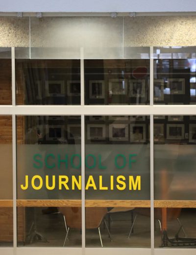 A photo of the glass outside of the J-school reads “SCHOOL OF JOURNALISM” in green and yellow, with photographs of past Minifie lecturers vaguely seen on the wall, blurred by the glass.