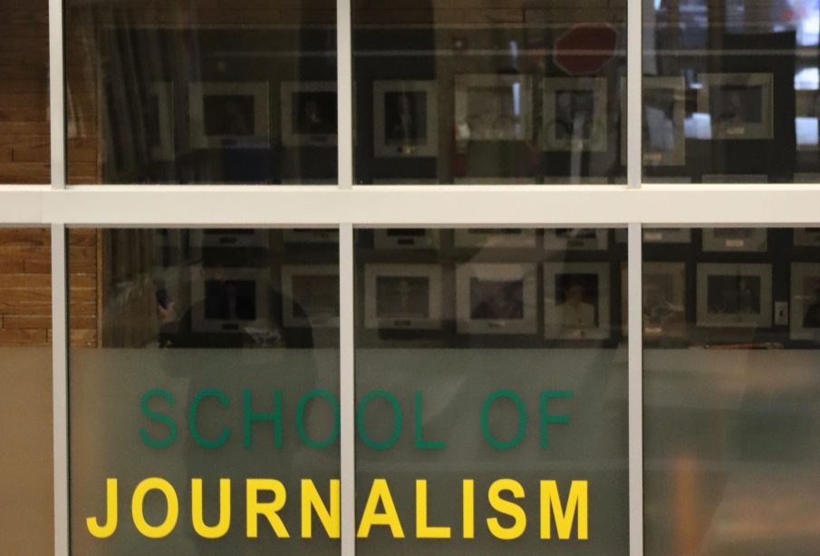 A photo of the glass outside of the J-school reads “SCHOOL OF JOURNALISM” in green and yellow, with photographs of past Minifie lecturers vaguely seen on the wall, blurred by the glass.