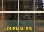 A photo of the glass outside of the J-school reads “SCHOOL OF JOURNALISM” in green and yellow, with photographs of past Minifie lecturers vaguely seen on the wall, blurred by the glass.