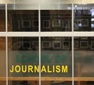 A photo of the glass outside of the J-school reads “SCHOOL OF JOURNALISM” in green and yellow, with photographs of past Minifie lecturers vaguely seen on the wall, blurred by the glass.