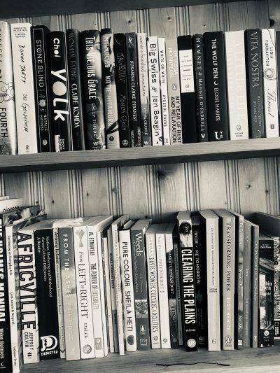 A black and white photograph of a bookshelf with two full shelves. The top shelf includes Thrust by Lidia Yuknavitch; Vita Nostra by Marina and Sergey Dyachenko; A Little Life by Hanya Yanaghihara; The Wolf Den by Elodie Harper; Hamnet by Maggie O’Farrell; The Secret History by Donna Tartt; My Year of Rest and Relaxation by Ottessa Moshfegh; White on White by Ayşegül Savaş; Big Swiss by Jen Beagin; Across a Hundred Mountains by Reyna Grande; Piranesi by Susanna Clarke; Jawbone by Mónica Ojeda; Intimacies by Katie Kitamura; This Vicious Grace by Emily Thiede; Woman, Eating by Claire Kohida; Yolk by Mary H.K. Choi; Stone Blend by Natalie Haynes; The Goldfinch by Donna Tartt; and Fourth Wing by Rebecca Yarros. The second shelf holds Gate 7 by manga artist group Clamp; Cabin Porn by Little, Brown and Company; Chromophobia by David Batchelor; Summer 2020 Ecology Issue No. 41 of Qwerty; Spring 2023 Issue No. 295 of The Fiddlehead; Transforming Power by Judy Rebick; Determinants of Indigenous Peoples’ Health Second Edition by Greenwood, de Leeuw, and Lindsay; Me Tomorrow: Indigenous Views of the Future by Douglas & McIntyre; Clearing The Plains by James William Daschuk; The Communist Manifesto by Marx and Engels; Saskatchewan Dirt: A Pandemic Quest for Connection by Bev Lundahl; Survival of the Friendliest by Brian Hare and Vanessa Woods; Women, Culture, & Politics by Angela Y. Davis; Weeds of Canada and the Northern United States by Royer & Dickinson; Pure Colour by Sheila Heti; Listening to the Beat of our Drum edited by Carrie Bourass, Elder Betty McKenna and Darlene Juschka; Noticed, Known and Missed by Cara Milne; The Power of Disability by Al Etmanski; and From Left to Right: Saskatchewan’s Political and Economic Transformation by Dale Eisler.