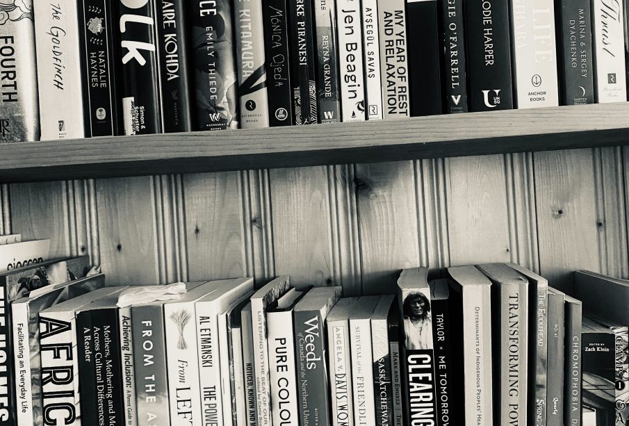 A black and white photograph of a bookshelf with two full shelves. The top shelf includes Thrust by Lidia Yuknavitch; Vita Nostra by Marina and Sergey Dyachenko; A Little Life by Hanya Yanaghihara; The Wolf Den by Elodie Harper; Hamnet by Maggie O’Farrell; The Secret History by Donna Tartt; My Year of Rest and Relaxation by Ottessa Moshfegh; White on White by Ayşegül Savaş; Big Swiss by Jen Beagin; Across a Hundred Mountains by Reyna Grande; Piranesi by Susanna Clarke; Jawbone by Mónica Ojeda; Intimacies by Katie Kitamura; This Vicious Grace by Emily Thiede; Woman, Eating by Claire Kohida; Yolk by Mary H.K. Choi; Stone Blend by Natalie Haynes; The Goldfinch by Donna Tartt; and Fourth Wing by Rebecca Yarros. The second shelf holds Gate 7 by manga artist group Clamp; Cabin Porn by Little, Brown and Company; Chromophobia by David Batchelor; Summer 2020 Ecology Issue No. 41 of Qwerty; Spring 2023 Issue No. 295 of The Fiddlehead; Transforming Power by Judy Rebick; Determinants of Indigenous Peoples’ Health Second Edition by Greenwood, de Leeuw, and Lindsay; Me Tomorrow: Indigenous Views of the Future by Douglas & McIntyre; Clearing The Plains by James William Daschuk; The Communist Manifesto by Marx and Engels; Saskatchewan Dirt: A Pandemic Quest for Connection by Bev Lundahl; Survival of the Friendliest by Brian Hare and Vanessa Woods; Women, Culture, & Politics by Angela Y. Davis; Weeds of Canada and the Northern United States by Royer & Dickinson; Pure Colour by Sheila Heti; Listening to the Beat of our Drum edited by Carrie Bourass, Elder Betty McKenna and Darlene Juschka; Noticed, Known and Missed by Cara Milne; The Power of Disability by Al Etmanski; and From Left to Right: Saskatchewan’s Political and Economic Transformation by Dale Eisler.