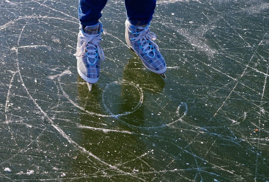 A photo of someone standing on ice in ice skates. The ice is scratched up and may be a frozen lake. Only the person’s skates and jeans are visible.