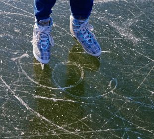 A photo of someone standing on ice in ice skates. The ice is scratched up and may be a frozen lake. Only the person’s skates and jeans are visible.