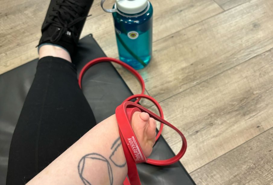 A photo of shae sackman holding a red resistance band. A blue water bottle is on the floor, and sackman sits on a black mat.