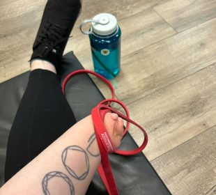 A photo of shae sackman holding a red resistance band. A blue water bottle is on the floor, and sackman sits on a black mat.
