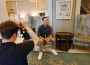 A photo of a person sitting on a chair being filmed by another person on a TV camera.