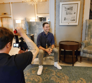 A photo of a person sitting on a chair being filmed by another person on a TV camera.