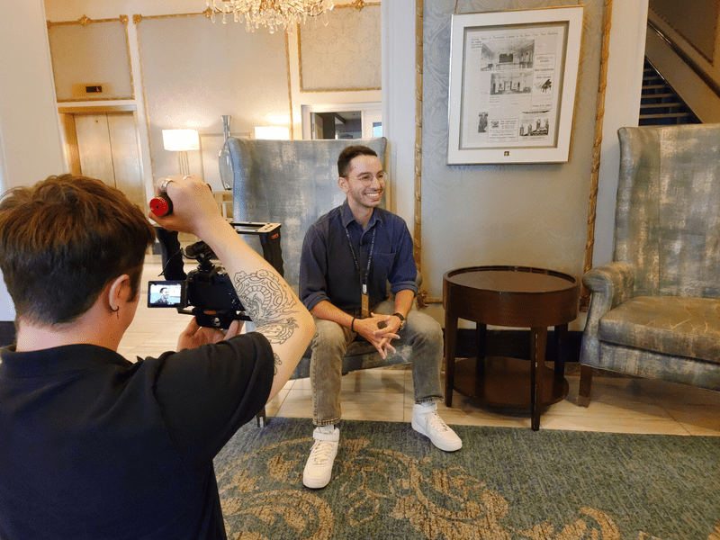 A photo of a person sitting on a chair being filmed by another person on a TV camera.