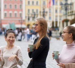 A photograph of a group of three people in business casual wear standing in a public space and joking with each other while two are holding coffees in one hand. The one on the right has a jacket draped over their arm.  