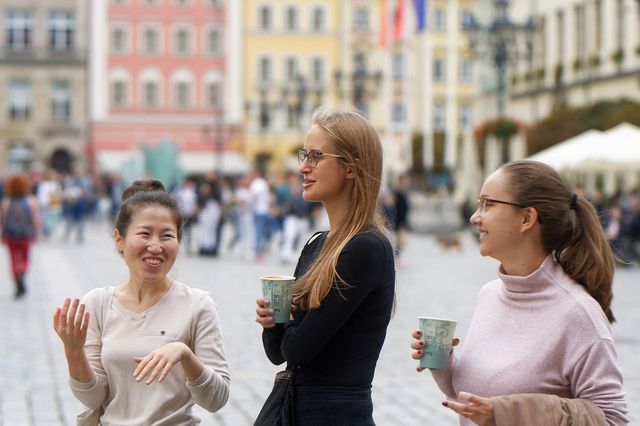 A photograph of a group of three people in business casual wear standing in a public space and joking with each other while two are holding coffees in one hand. The one on the right has a jacket draped over their arm.  