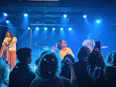 A photograph of Hotel Mira performing at Louis' in Saskatoon on February 1, 2024. The photo was taken in the section closest to the stage where people stand to watch the show, and there are other show attendees also taking photos or videos in the shot.