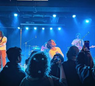A photograph of Hotel Mira performing at Louis' in Saskatoon on February 1, 2024. The photo was taken in the section closest to the stage where people stand to watch the show, and there are other show attendees also taking photos or videos in the shot.