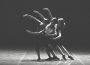 A black and white photograph of three ballet dancers performing under a spotlight. They have their right arm raised and the other bent at their waist, and are leaning slightly to their left with their left leg extended to the side.