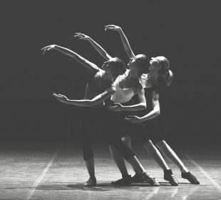 A black and white photograph of three ballet dancers performing under a spotlight. They have their right arm raised and the other bent at their waist, and are leaning slightly to their left with their left leg extended to the side.