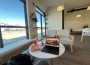 A café table with a notebook and laptop on the desk, right next to a window with the view of a bright sunny day.