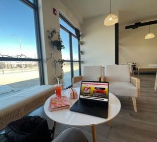 A café table with a notebook and laptop on the desk, right next to a window with the view of a bright sunny day.