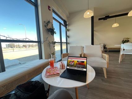 A café table with a notebook and laptop on the desk, right next to a window with the view of a bright sunny day.