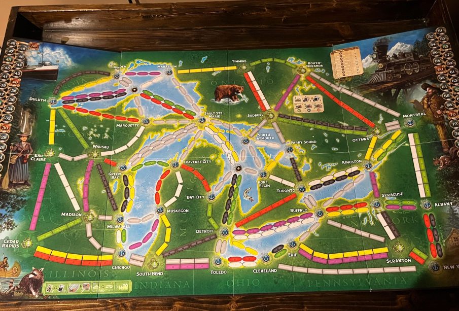 A photograph of a board for a board game. The board is covered in spaces for trains to be placed and images of characters or animals, and has the background of a coloured map of an area in southern Ontario and some northern states beneath it.