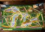 A photograph of a board for a board game. The board is covered in spaces for trains to be placed and images of characters or animals, and has the background of a coloured map of an area in southern Ontario and some northern states beneath it.