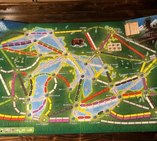 A photograph of a board for a board game. The board is covered in spaces for trains to be placed and images of characters or animals, and has the background of a coloured map of an area in southern Ontario and some northern states beneath it.