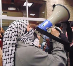 A photograph of a protester wearing a keffiyeh and speaking into a megaphone while walking in a group.