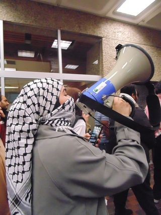 A photograph of a protester wearing a keffiyeh and speaking into a megaphone while walking in a group.