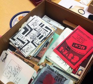 A cardboard box filled with books, pamphlets, and zines of all colours.