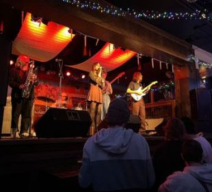 A photograph of a band playing on stage, taken from the left side of the standing section of the audience.