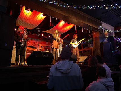 A photograph of a band playing on stage, taken from the left side of the standing section of the audience.