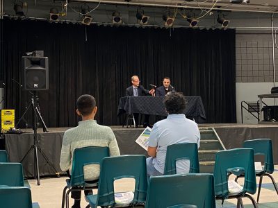 A photo is taken from behind the front row at a town hall with university executives. Jeff Keshen is pictured at a table on the stage.