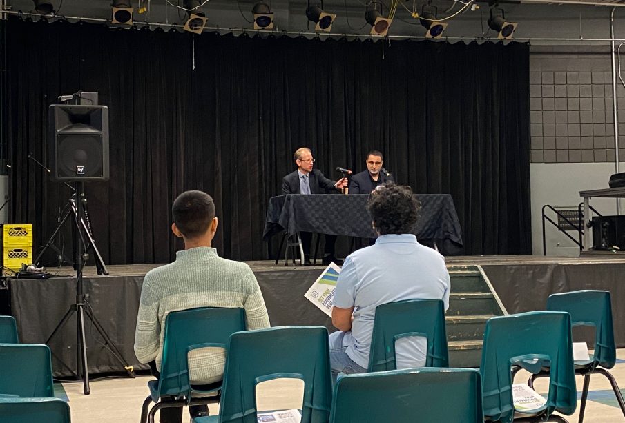 A photo is taken from behind the front row at a town hall with university executives. Jeff Keshen is pictured at a table on the stage.