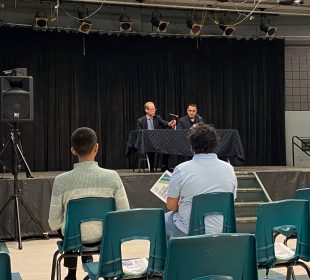 A photo is taken from behind the front row at a town hall with university executives. Jeff Keshen is pictured at a table on the stage.