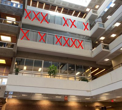 The windows of second, third, and fourth floor faculty offices in the Administration-Humanities building are seen from below. Large red “X”s cross out many of the offices