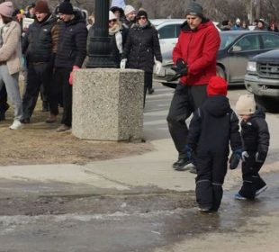 Teachers walk in a large group in front of the Saskatchewan Legislature. In front of them, just off of the sidewalk, two children stand on ice.