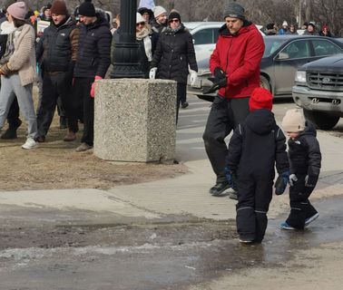Teachers walk in a large group in front of the Saskatchewan Legislature. In front of them, just off of the sidewalk, two children stand on ice.