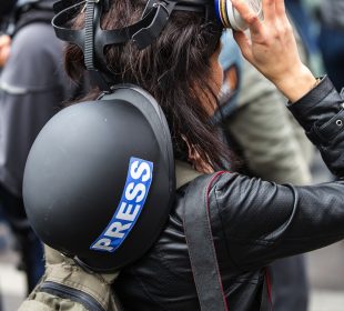 A faceless journalist is pictured from behind. They have a helmet reading “PRESS” slung across their back and have lifted respiratory protection up from their face. Their hand rests on the mask.