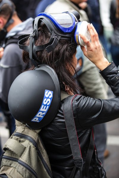 A faceless journalist is pictured from behind. They have a helmet reading “PRESS” slung across their back and have lifted respiratory protection up from their face. Their hand rests on the mask.