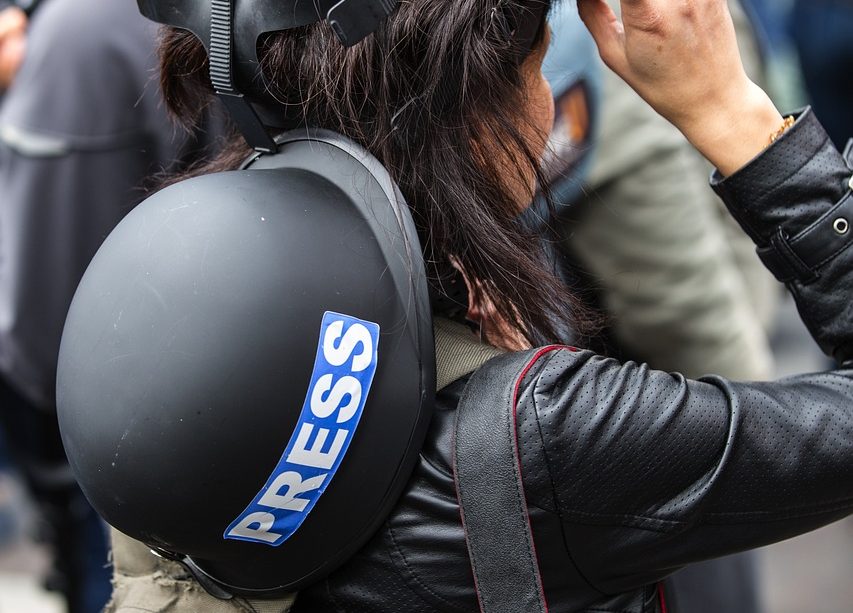 A faceless journalist is pictured from behind. They have a helmet reading “PRESS” slung across their back and have lifted respiratory protection up from their face. Their hand rests on the mask.