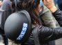 A faceless journalist is pictured from behind. They have a helmet reading “PRESS” slung across their back and have lifted respiratory protection up from their face. Their hand rests on the mask.