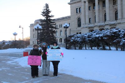 Three people standing with signs/placards at an STF rally in front of the legislative building.
