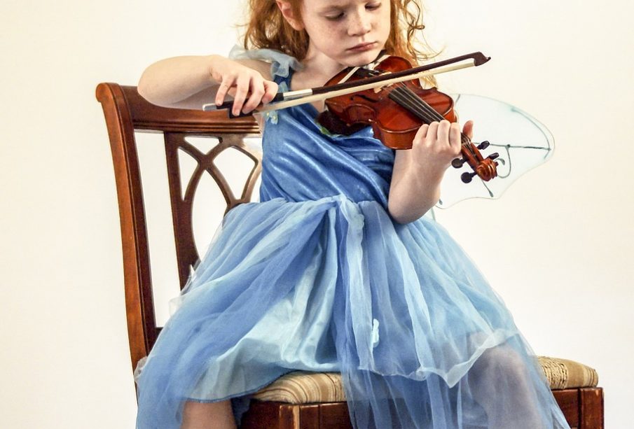 A young child in a blue dress playing the violin while sitting on a wooden chair.