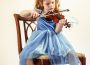 A young child in a blue dress playing the violin while sitting on a wooden chair.