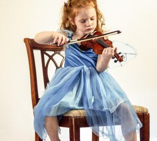 A young child in a blue dress playing the violin while sitting on a wooden chair.