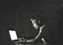 A person sitting in a dark room has their face illuminated by a laptop screen. They are typing intently.