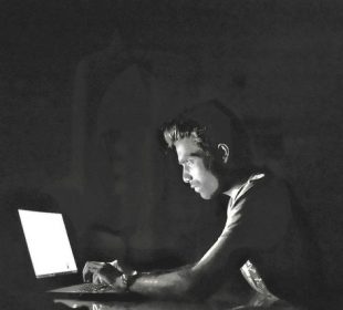 A person sitting in a dark room has their face illuminated by a laptop screen. They are typing intently.